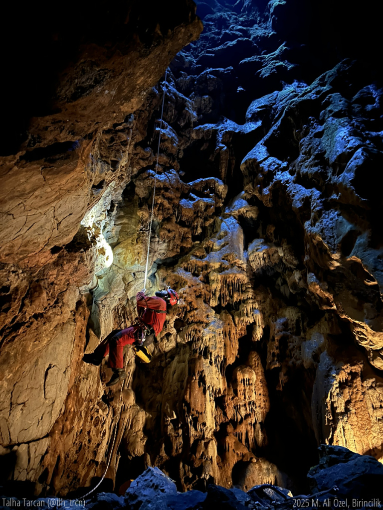 İZMAD’ın ilk Kurcuk Düdeni faaliyetinde, ben (Talha Tarcan) ve Görkem Doğan ipten süzülerek mağaraya indik. Aşağı vardığımızda karşımızda öyle bir manzara vardı ki, sanki National Geographic setine yanlışlıkla dalmış gibiydik. Döşeme ekibi işi abartmış; mağaranın dibine LED ışık yerleştirmiş. Yukarıdan süzülen gün ışığıyla birleşince ortaya tam bir ‘yeraltı şovu’ çıkmıştı. Toz zerrecikleri bile ışığın içinde ağır çekimde dans ediyordu. Biz manzaranın büyüsüne kapılmışken Görkem efsane cümlesini patlattı: “En iyi fotoğrafı çekene 50 TL!” Bir anda mağara keşif alanından çıkıp yeraltı fotoğraf yarışması setine döndü. Herkes en iyi açı, en iyi ışık peşinde… O an sadece biz, ışık ve o 50 TL’lik şan vardı. Tabii ki Görkem o 50 TL’yi kimseye vermedi. Ama ne fark eder? Çünkü o an — ışığın, sessizliğin ve biraz da deliliğin birleştiği o kare — paradan çok daha değerliydi. Sonuç? Ödül yok. Ama anı? Efsane.